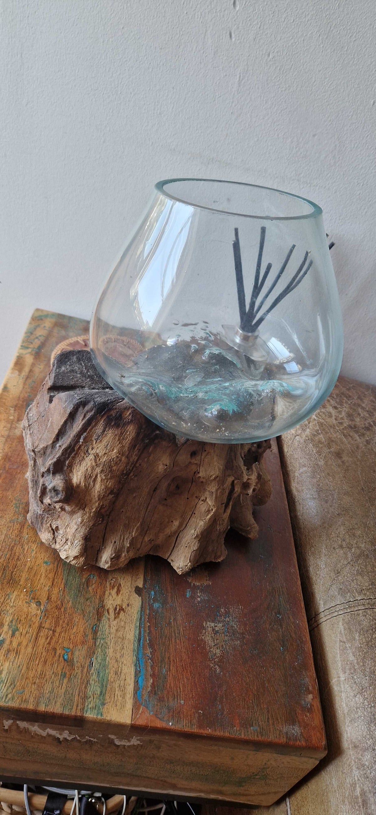 A clear glass bowl-shaped vase or planter with a black reed diffuser stick inside, displayed on a rustic piece of driftwood resting on a wooden box against a plain wall.