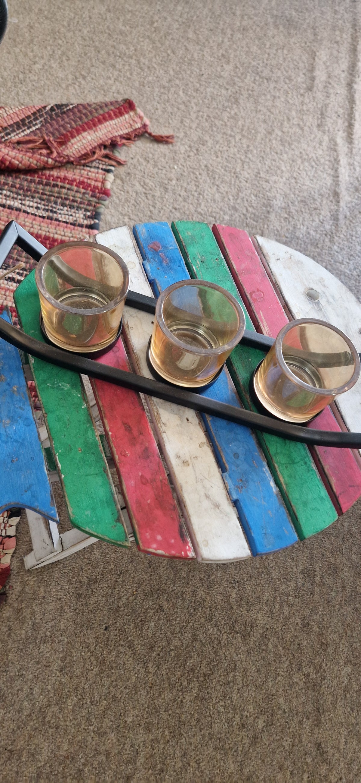 A colorful round wooden slat tray holding three clear drinking glasses with amber liquid, placed on a textured beige carpet with a patterned textile visible at the lower left.